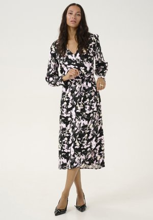 Robe noire ajustée avec un motif abstrait rose et blanc, manches longues, taille cintrée et jupe fluide. Portée avec des escarpins noirs à bouts pointus.