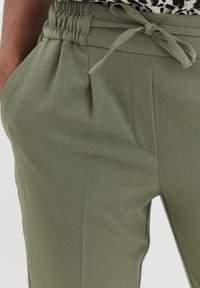 Pantalon vert clair avec une taille froncée, cordon de serrage élastique et poches latérales ; texture lisse, coupe ajustée, avec plis avant.