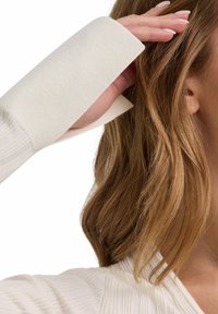 Langsleeve, geribbelde top in off-white, met brede manchetten. De stof lijkt zacht, met een subtiele textuur en gedetailleerd stikwerk.