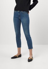Jeans ajustados de color azul con una cintura media, largo hasta el tobillo y diseño clásico de cinco bolsillos. Combinados con mocasines negros que presentan un acento de cadena dorada.