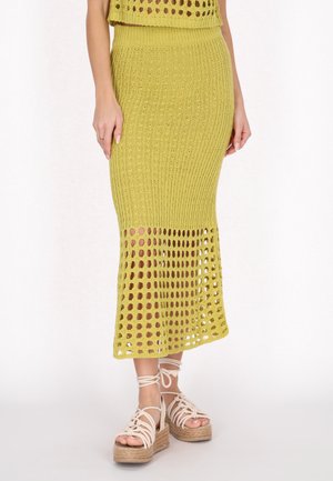 Vrouw draagt een gele gebreide midi-rok met open geweven zoom en beige platformsandalen met enkelbandjes op een witte achtergrond.