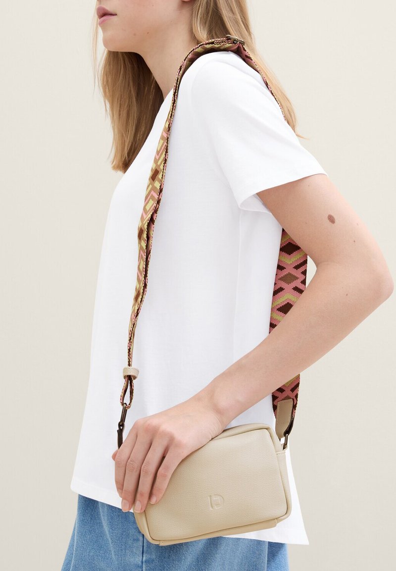 Borsa a tracolla in pelle beige con una tracolla in tessuto motivo geometrico nei colori rosa e marrone, tenuta da una persona in camicia bianca.