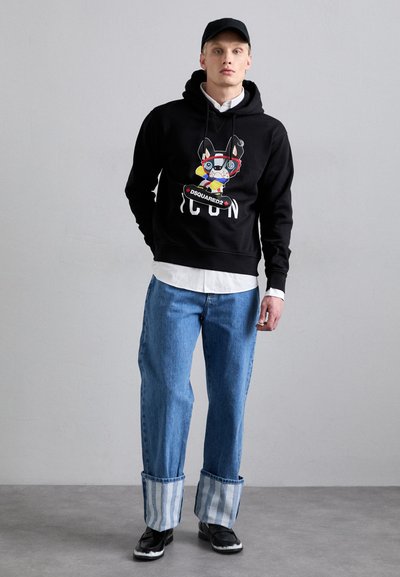 Svart hoodie med grafisk design och texten "ICON", tillsammans med lösa blå denimjeans med randiga muddar och svarta skor.