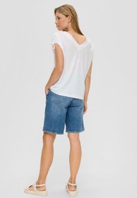 T-shirt bianco, con un taglio rilassato, scollo a V e maniche arrotolate, abbinato a pantaloni corti in denim blu, con orlo sfilacciato. Sandali chiari completano l'outfit.