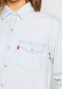 Light blue denim shirt featuring a button-up design, pointed collar, and a single chest pocket with a contrasting red logo tag.