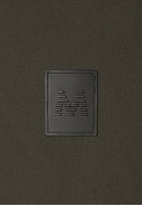 Svart fyrkantig läderlapp med präglade horisontella linjer som bildar bokstaven M på en mörk texturerad tygbakgrund.