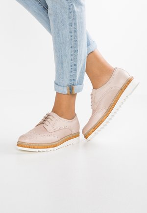 Hellrosa Brogue-Schuhe aus Leder mit weißen gezackten Sohlen, getragen zu hochgekrempelten hellblauen Jeans auf weißem Boden.