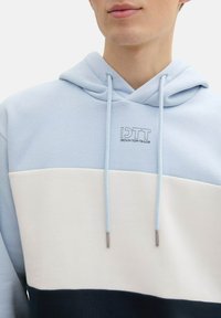 Bluza z kapturem w kolorze jasnoniebieskim na górze, białym w środkowej części oraz granatowym na dolnej części. Zawiera logo oraz sznurki ze metalowymi końcówkami.