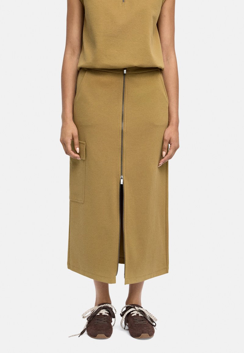 Oliwkowa, fakturowana bawełniana spódnica z przednim zamkiem, bocznymi kieszeniami i prostym krojem, zestawiona z brązowymi sneakersami i sznurówkami.