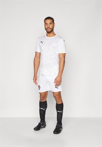 Hvid PUMA fodboldtrøje med subtil mønstret tekstur, sort logo på brystet, parret med hvide shorts og sorte knæstrømper.