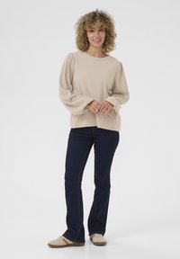 Beige langærmet top med voluminøse, ribbede ballonærmer. Sat sammen med mørkeblå flagrende jeans og beige sneakers med brune accenter.