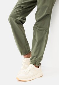 Beige sportieve sneakers met een gestructureerde bovenkant en witte veters, gecombineerd met losse, olijfgroene broek met elastische boorden.