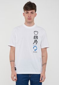 T-shirt bianca a maniche corte in cotone. Presenta testo stampato in nero e blu e un logo sulla parte anteriore, con un piccolo etichetta di lato.