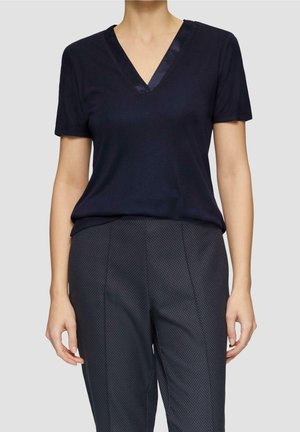 Kobieta w krótkim rękawie czarnej koszulce z dekoltem w serek i ciemnych spodniach w wzory, stojąca na jednolitym, szarym tle.