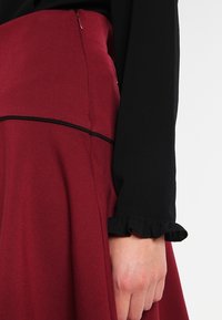 Jupe bordeaux avec une texture lisse, dotée d'une bordure noire le long de la taille. Un haut noir avec des poignets froncés est partiellement visible.