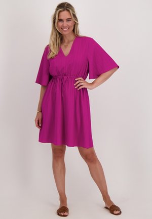Korte roze jurk met een V-hals, korte batwing-mouwen en een verstelbare trekkoord in de taille. Glad stof met een subtiele textuur. Bruine platte sandalen.