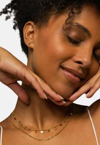 Goldbeschichtete Halsketten mit runden Anhängern, zarten Kettenlinks und einer feinen Textur. Das Model posiert mit den Händen unter dem Kinn.