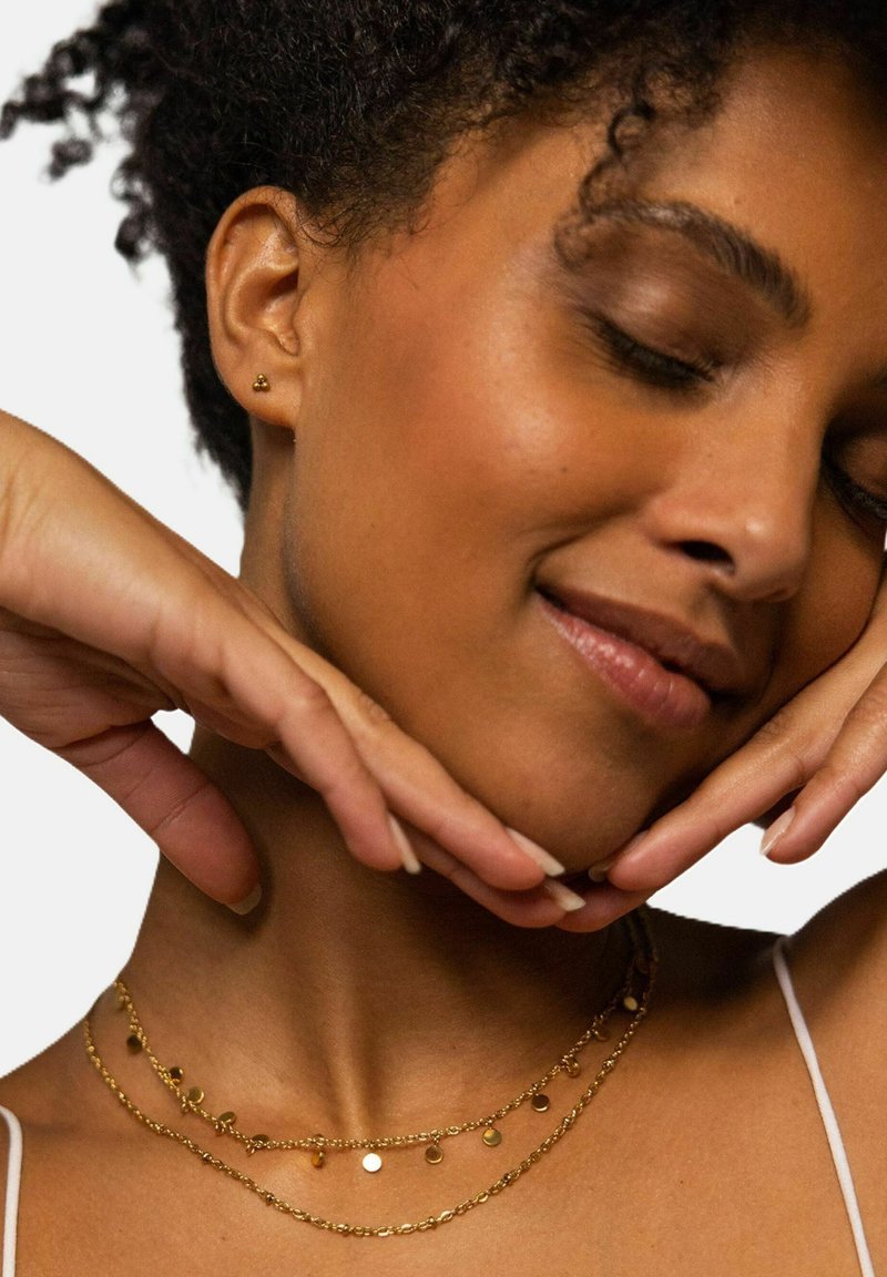 Goldbeschichtete Halsketten mit runden Anhängern, zarten Kettenlinks und einer feinen Textur. Das Model posiert mit den Händen unter dem Kinn.