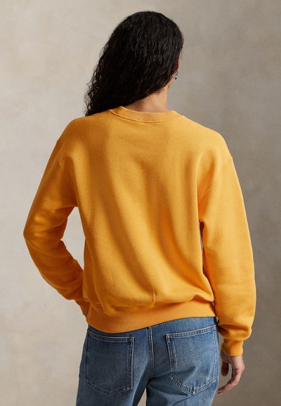 Żółta bluza z okrągłym dekoltem wykonana z miękkiego materiału, z prążkowanymi mankietami i dolnym obszyciem, zestawiona z niebieskimi dżinsami o luźnym kroju.