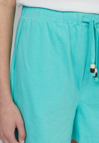 Pantaloni shorts turchesi realizzati in un tessuto morbido, con vita elastica e cordoncino decorato con perline di legno.