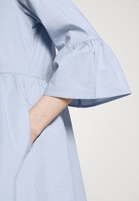 Hellblaues Kleid mit weit ausgeflogenen Ärmeln, strukturiertem Stoff und seitlichen Taschen. Das Design umfasst eine geraffte Taille und eine glatte Oberfläche.