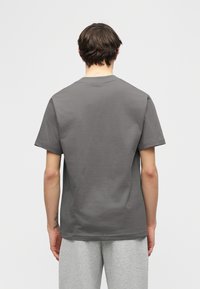 Άτομο με κοντά καστανά μαλλιά φορώντας απλό σκούρο γκρι κοντομάνικο t-shirt και ανοιχτό γκρι παντελόνι, στέκεται με την πλάτη προς τα εμπρός σε ουδέτερο φόντο.