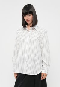 Camicia bianca con righe verticali nere, dotata di colletto a punta e maniche lunghe con polsini striati. Tessuto liscio, vestibilità comoda.