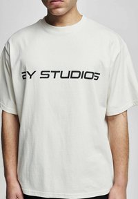 Valkoinen puuvillapaita, jossa on musta painettu teksti "2Y STUDIOS" edessä. Rentoa tyyliä, lyhyet hihat.
