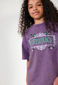 Lila, överdimensionerad t-shirt med en grafisk design som visar ordet "BEETLEJUICE" i grönt och ormar, korta ärmar och en mjuk textur.
