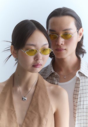 Lunettes de soleil teintées de jaune avec de fines montures en argent, associées à un haut sans manches beige, entourées d'un arrière-plan flou.