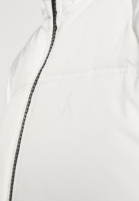 Dettaglio ravvicinato di una giacca bianca con zip nera e un logo "cK" in rilievo discreto nella zona del petto.