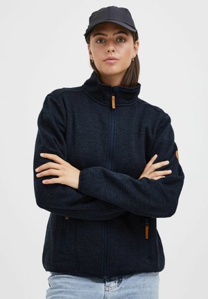 Fleecejacke in Dunkelnavy mit hochschließendem Kragen, Reißverschluss und Seitentaschen. Ausgestattet mit strukturiertem Stoff und kontrastierendem, hellbraunem Reißverschlussschieber.