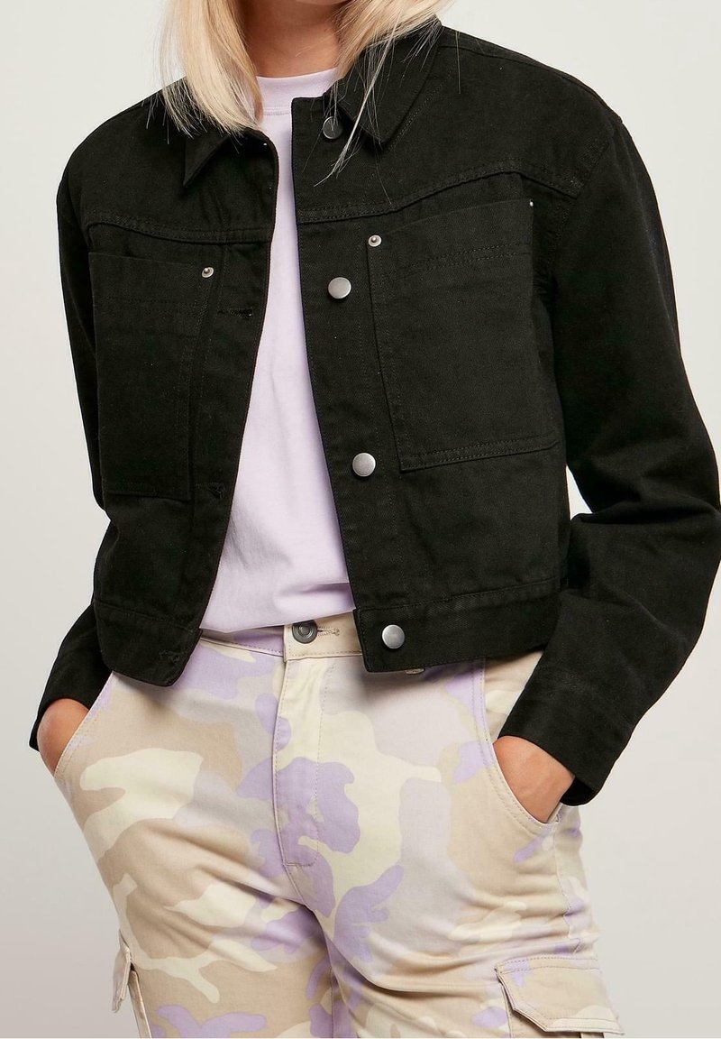 Svart denimkorta jacka med silverknappar, två bröstfickor och krage, bärs över en ljuslila topp och mönstrade beige byxor.