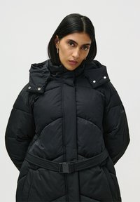 Mujer con cabello negro lacio, vistiendo un abrigo de plumas negro ceñido con un cinturón, con un cuello alto y pendientes de aro plateados, contra un fondo liso.