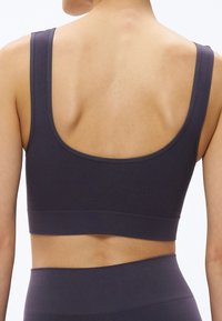 Žena v tmavo fialovej športovej podprsenke a zodpovedajúcich legínach s vysokým pásom, zobrazená zozadu na jednoduchom svetlom pozadí.