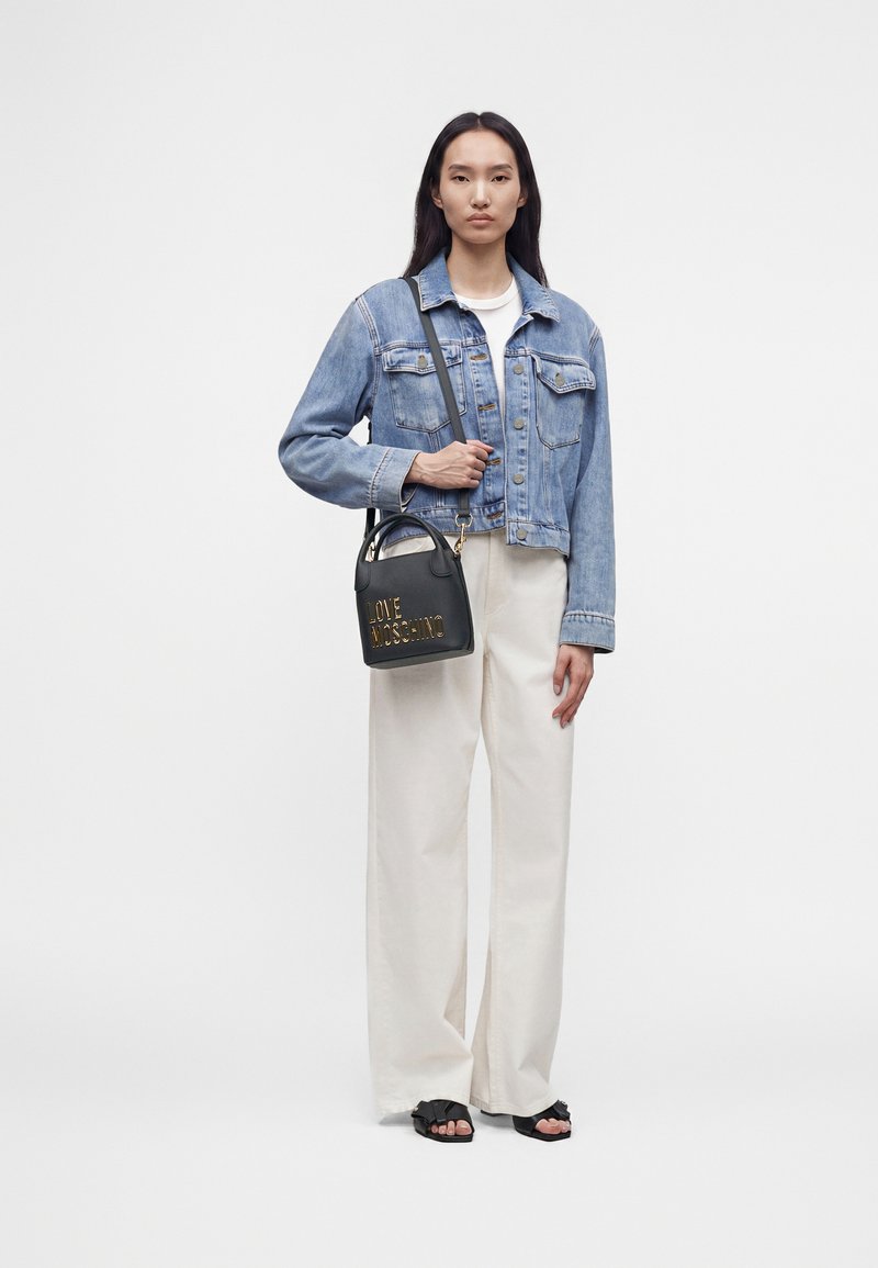 Γυναίκα με ανοιχτό τζιν μπουφάν, λευκό παντελόνι με φαρδιά παντελόνια, μαύρα σανδάλια, κρατώντας μαύρη τσάντα Love Moschino με χρυσά γράμματα.