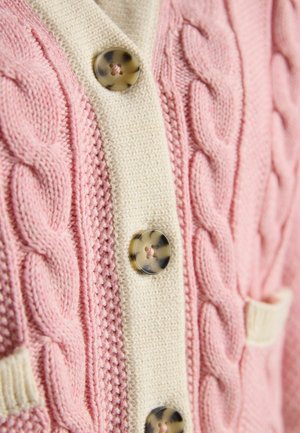 Nahaufnahme eines pinkfarbenen, kabelgestrickten Cardigans mit cremefarbenem Knopfleistenbesatz und Schildpattknöpfen.