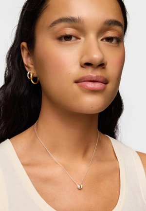 Giovane donna con lunghi capelli scuri che indossa piccoli orecchini a cerchio in argento e oro e una collana con pendente a cuore bicolore su una catena in argento.