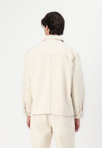 Lys beige denimjakke med standard krave, lange ærmer og knapmanchetter. Har syningsdetaljer og et lige snitdesign.