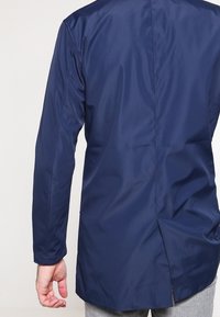 Námořnická modrá bunda s dlouhým rukávem a hladkým, matným povrchem. Obsahuje zadní rozparek a minimální švy. Žádné viditelné kování.