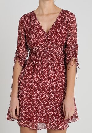 Rode bloemenjurk met V-hals, gerimpelde driekwart mouwen gebonden met strikken, en een geplooide taille, gedragen door een staand persoon.