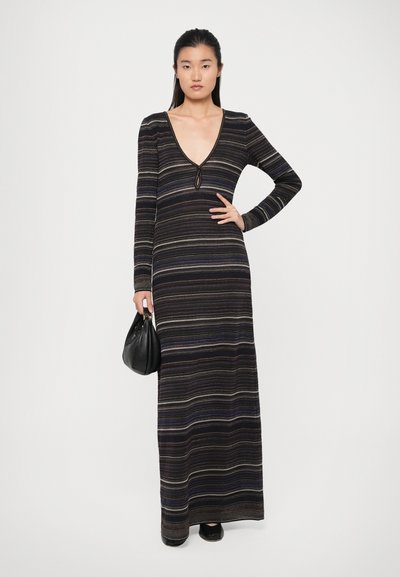 Abito maxi a righe in tonalità scure con maniche lunghe, scollatura a V e dettaglio a oblò. Il modello tiene una borsa nera, mostrando una silhouette aderente.
