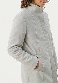 Hellgrauer Wollmantel mit hohem Kragen, seitlichen Taschen und subtilen vertikalen Nahtdetails. Glatte Textur und figurbetonter Schnitt.