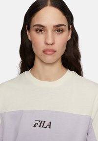 Väriestetty puuvillainen t-paita, jossa on vaalea keltainen yläosa, vaaleanlila alaosa ja musta FILA-logo edessä.