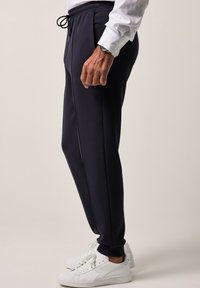 Granatowe dresy sportowe z sznurkiem w talii i kieszeniami bocznymi, charakteryzujące się dopasowanym krojem oraz kontrastowymi paskami wzdłuż szwów. W zestawieniu z białymi trampkami.