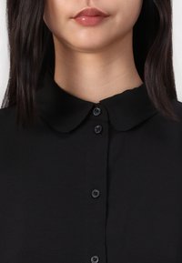 Black collared blouse with a button-down front, featuring round black buttons and a smooth texture. Minimalistic design, no patterns.