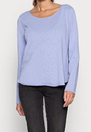 Mujer con una camiseta de manga larga azul claro y pantalones negros, de pie con los brazos relajados a los lados sobre un fondo sencillo.