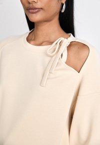 Sudadera beige con cuello redondo, que presenta un corte decorativo en el hombro y un detalle de lazo. Tela suave con una textura lisa.