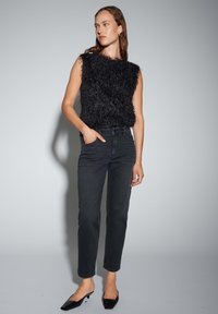 Femme debout portant un haut noir sans manches en tissu duveteux, un jean noir taille haute et des chaussures noires pointues à talons bas, regardant sur le côté.