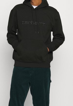 Person trägt einen schwarzen Carhartt Hoodie mit Fronttasche und dunkelgrünen Cordhosen vor einfarbigem Hintergrund.